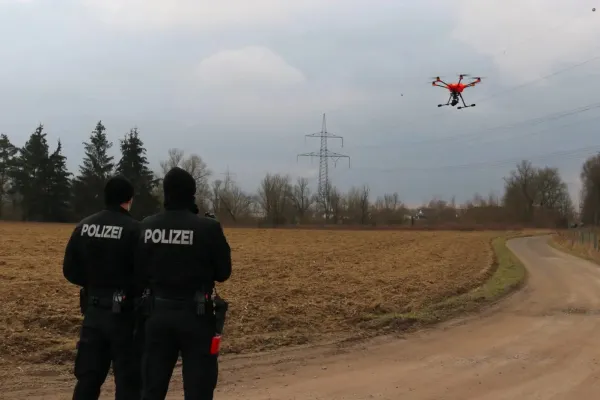 Zwei Polizist*innen stehen vor einem gepflügten Feld, vor ihnen fliegt ein Multicopter/Drohne.